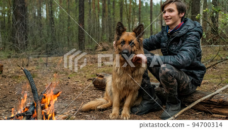 Young man with big dog sits at the fire in the forest. Cheerful guy is stroking his dog, outdoor. Happy caucasian teenager with his German shepherd by the fire in nature, in autumn. 94700314