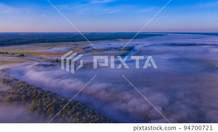 Aerial view of a beautiful summer landscape with a fog while dawn. Photo from drone of a foggy landscape in spring. Top view to land while sunrise. Early morning in nature, misty weather. Aerial view of a beautiful summer landscape with a fog while dawn. Photo from drone of a foggy landscape in spring. Top view to land while sunrise. Early morning in nature, misty weather. 94700721