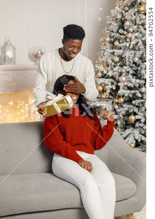 Young black couple with gifts in Christmas eve. Woman sitting on a coach at home. Man standing behind and presented a gift for his girlfriend. 94702554
