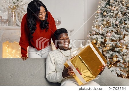 Young black couple with gifts in Christmas eve. Man sitting on a coach at home. Brunette woman standing behind and presented a gift for her boyfriend. Young black couple with gifts in Christmas eve. Man sitting on a coach at home. Brunette woman standing behind and presented a gift for her boyfriend. 94702564