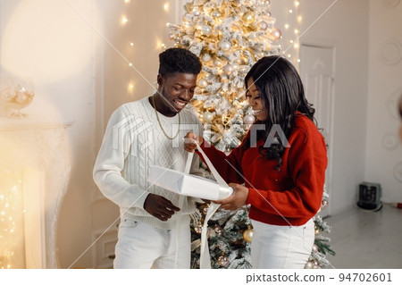 Happy black couple holding gift and standing near Christmas tree. Man amd woman celebrating winter holidays at home. Brunette woman wearing a red sweater. 94702601