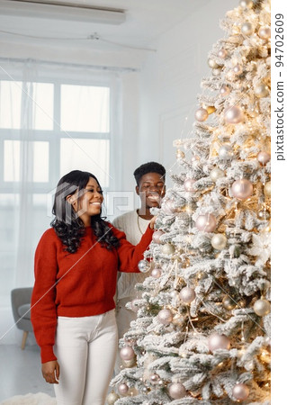 Happy black couple decorating Xmas tree together. Man amd woman celebrating winter holidays at home. Brunette woman wearing a red sweater. Happy black couple decorating Xmas tree together. Man amd woman celebrating winter holidays at home. Brunette woman wearing a red sweater. 94702609