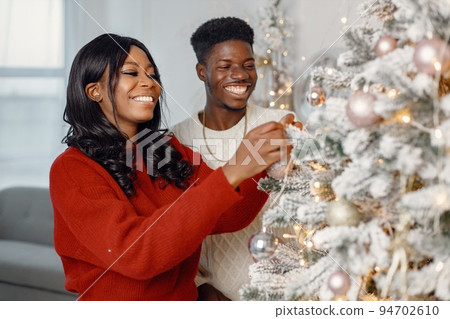 Happy black couple decorating Xmas tree together. Man amd woman celebrating winter holidays at home. Brunette woman wearing a red sweater. Happy black couple decorating Xmas tree together. Man amd woman celebrating winter holidays at home. Brunette woman wearing a red sweater. 94702610