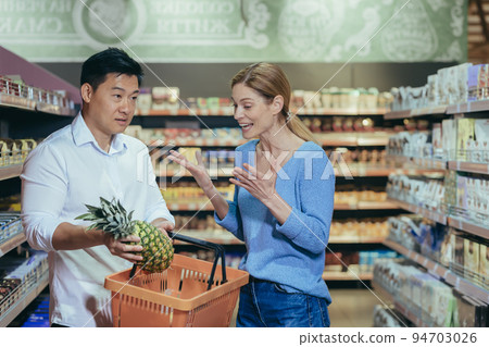 Family conflict in the store. Young interracial couple arguing in a supermarket. A woman shouts at an Asian man who spent money on expensive fruit. A man stands confused with a basket in his hands. Family conflict in the store. Young interracial couple arguing in a supermarket. A woman shouts at an Asian man who spent money on expensive fruit. A man stands confused with a basket in his hands. 94703026
