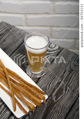 Thin sausages for beer. On a rectangular plate. Next to it is a glass of wheat beer. Background from pine boards. Close-up. 94703131