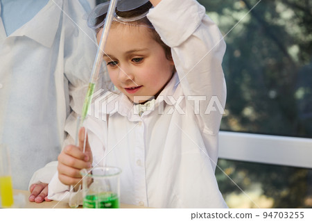 Smart Caucasian schoolgirl, elementary age student wearing white lab coat and goggles, conducts a chemical experiment in school Chemistry laboratory. Education, kids entertainment and development 94703255