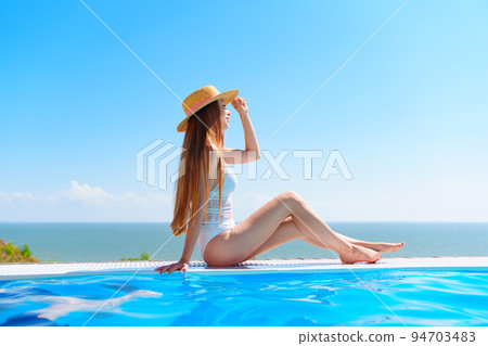 a slender woman in a white swimsuit in a straw hat sits on the pool. a slender woman in a white swimsuit in a straw hat sits on the pool. 94703483