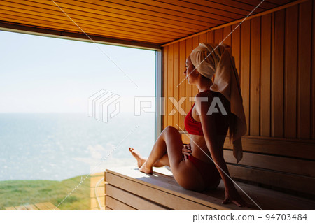 woman in a red swimsuit sits on a bench in the sauna with the panoramic window.  94703484