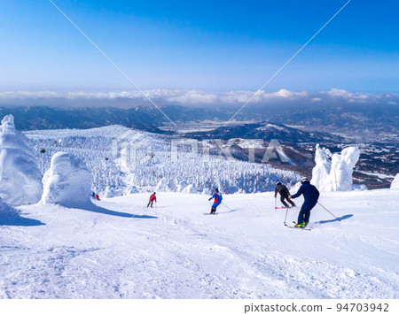 People skiing through a field of frost-covered trees at a sunny ski resort (Zao Onsen Ski Resort, Yamagata Prefecture) 94703942