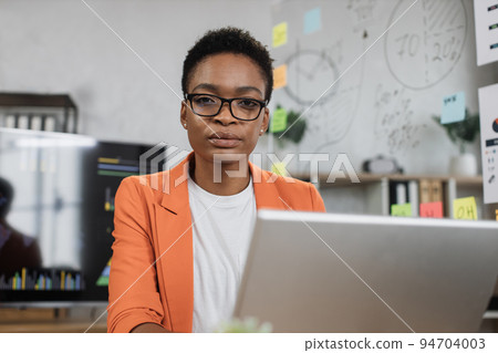 Portrait of african business woman in orange suit sitting at desk and looking at camera. Portrait of african business woman in orange suit sitting at desk and looking at camera. 94704003