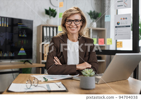 Beautiful senior woman with blond hair looking at camera while sitting at desk with papers 94704038