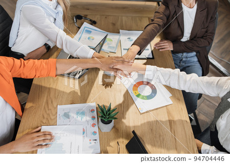 Top view of multiracial female business people stacking their hands together while sitting together Top view of multiracial female business people stacking their hands together while sitting together 94704446