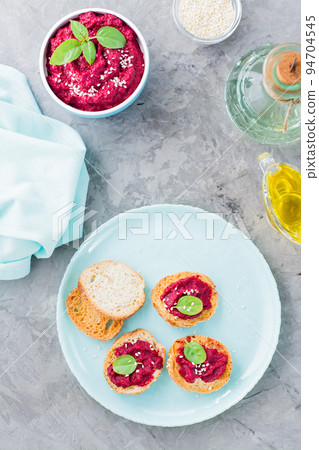 Homemade bruschetta with beetroot hummus on small baked baguette toast on a plate and a bowl of hummus on the table. Vertical and top view Homemade bruschetta with beetroot hummus on small baked baguette toast on a plate and a bowl of hummus on the table. Vertical and top view 94704545