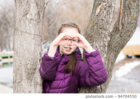 Young attractive girl in glasses makes a heart shape with her palms in front of her face in a city park in early spring 94704550
