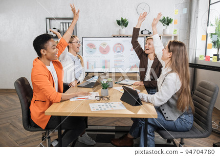 Female experts celebrating successful negotiation while sitting at desk and holding hands up 94704780
