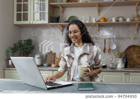 Young beautiful hispanic woman with curly hair working at home using laptop, woman in kitchen smiling and happy remote work freelancer. Young beautiful hispanic woman with curly hair working at home using laptop, woman in kitchen smiling and happy remote work freelancer. 94704950
