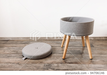 Trendy stool with and build-in storage space. Side view, white wall, copy space. Modern mulrifunctiional chair with wooden legs and removable lid. Round gray linen pouf 94706146