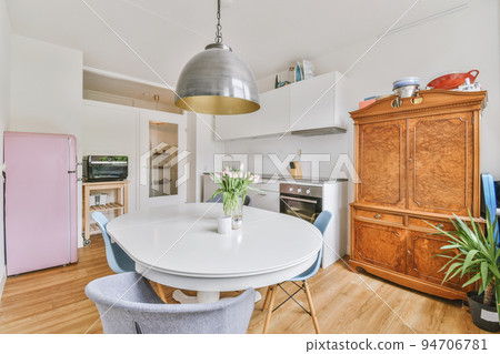 Modern minimalist style interior design of studio apartment with open white kitchen and dining zone with table and chairs illuminated by loft style lamp Modern minimalist style interior design of studio apartment with open white kitchen and dining zone with table and chairs illuminated by loft style lamp 94706781
