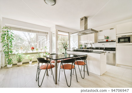 Modern minimalist style interior design of studio apartment with open white kitchen and dining zone with table and chairs illuminated by loft style lamp 94706947