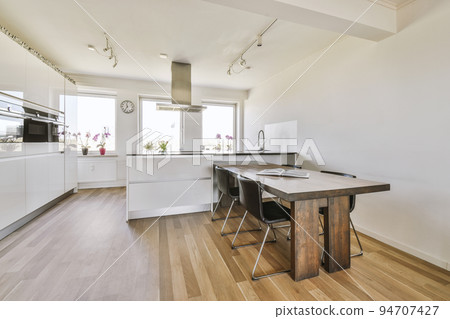 Contemporary minimalist style interior design of light studio apartment with wooden table and chairs in dining zone between open kitchen and living room with white walls and parquet floor 94707427