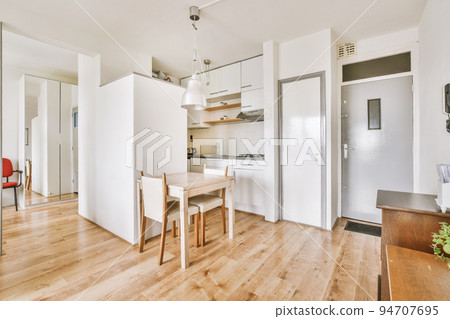 Contemporary minimalist style interior design of light studio apartment with wooden table and chairs in dining zone between open kitchen and living room with white walls and parquet floor 94707695