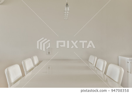 Modern minimalist style interior design of studio apartment with open white kitchen and dining zone with table and chairs illuminated by loft style lamp Modern minimalist style interior design of studio apartment with open white kitchen and dining zone with table and chairs illuminated by loft style lamp 94708358