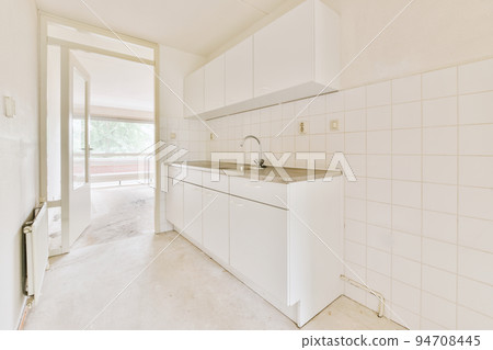 Fragment of interior of light narrow home kitchen with minimalist style white furniture with sink and stove Fragment of interior of light narrow home kitchen with minimalist style white furniture with sink and stove 94708445