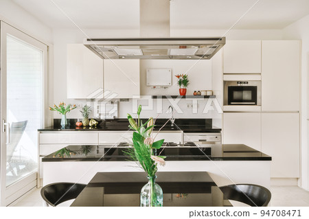 Modern minimalist style interior design of studio apartment with open white kitchen and dining zone with table and chairs illuminated by loft style lamp Modern minimalist style interior design of studio apartment with open white kitchen and dining zone with table and chairs illuminated by loft style lamp 94708471
