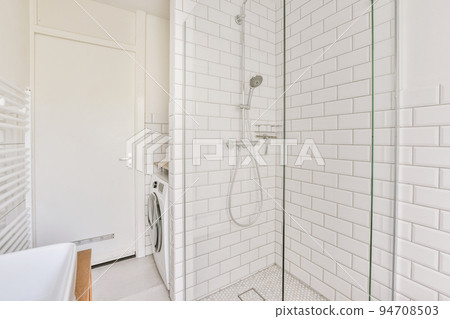 Sinks with mirrors and clean bathtub,washing machine located near shower box with glass door in modern bathroom with white tiled walls Sinks with mirrors and clean bathtub,washing machine located near shower box with glass door in modern bathroom with white tiled walls 94708503