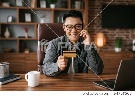 Glad middle aged korean man in glasses calls by phone use laptop and credit card, shopping online 94708506
