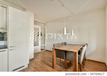 Modern minimalist style interior design of studio apartment with open white kitchen and dining zone with table and chairs illuminated by loft style lamp Modern minimalist style interior design of studio apartment with open white kitchen and dining zone with table and chairs illuminated by loft style lamp 94708627