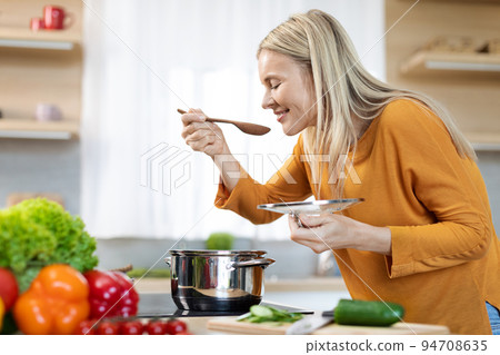 Pretty middle aged woman tasting food while cooking, kitchen interior 94708635