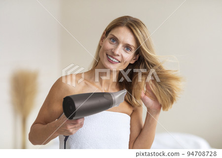Happy mature woman drying hair with hairdryer, standing wrapped in towel in bathroom at home after morning shower Happy mature woman drying hair with hairdryer, standing wrapped in towel in bathroom at home after morning shower 94708728