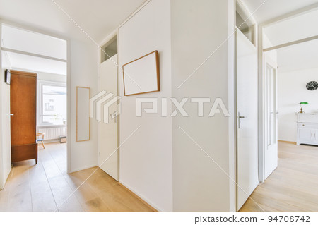 Narrow corridor with white walls and doors leading to spacious room with windows and parquet floor in modern apartment 94708742