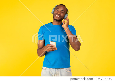African Guy Listening Music On Cellphone Wearing Earphones, Yellow Background African Guy Listening Music On Cellphone Wearing Earphones, Yellow Background 94708859