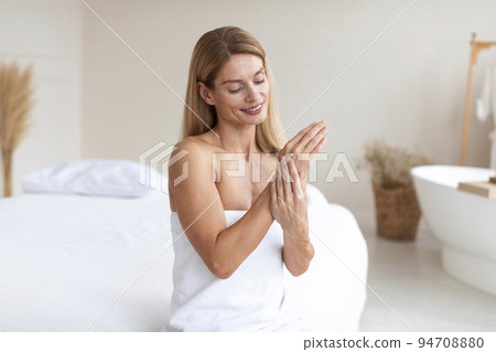 Satisfied middle aged lady in towel applying cream on hands, sitting on bed in bedroom interior, free space 94708880