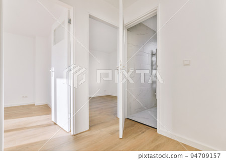 Narrow corridor with white walls and doors leading to spacious room with windows and parquet floor in modern apartment 94709157