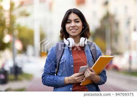 Modern Student. Smiling Young Arab Female With Workbooks And Smartphone Posing Outdoors Modern Student. Smiling Young Arab Female With Workbooks And Smartphone Posing Outdoors 94709375