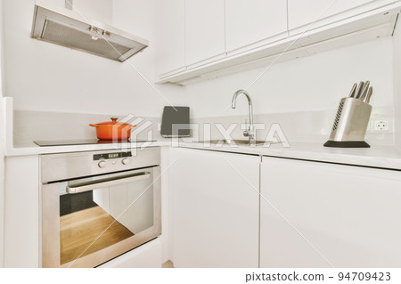Fragment of interior of light narrow home kitchen with minimalist style white furniture with sink and stove Fragment of interior of light narrow home kitchen with minimalist style white furniture with sink and stove 94709423