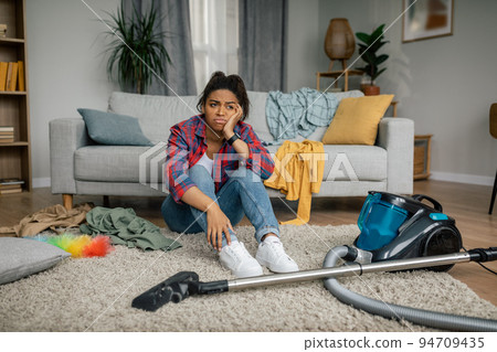 Unhappy tired young african american female sit on floor with vacuum cleaner, have problems with hygiene 94709435
