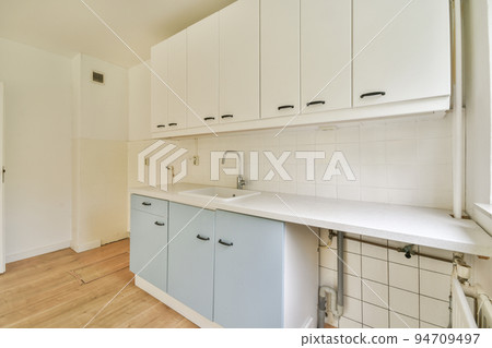 Fragment of interior of light narrow home kitchen with minimalist style white furniture with sink and stove Fragment of interior of light narrow home kitchen with minimalist style white furniture with sink and stove 94709497