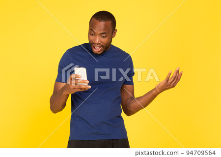 Discontented Black Male Athlete Holding Phone Over Yellow Studio Background Discontented Black Male Athlete Holding Phone Over Yellow Studio Background 94709564