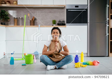 Glad young black lady sit on floor with mop, cleaning supplies meditating in white kitchen interior Glad young black lady sit on floor with mop, cleaning supplies meditating in white kitchen interior 94709621