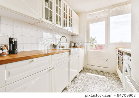 Fragment of interior of light narrow home kitchen with minimalist style white furniture with sink and stove Fragment of interior of light narrow home kitchen with minimalist style white furniture with sink and stove 94709908