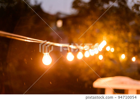 Decorative outdoor string lights hanging on the tree in the back yard at night time close, midges buzzing around on a summer evening 94710054