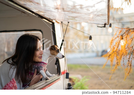 Caucasian woman looks out of the window of the van while hugging her dog Jack Russell Terrier. Travel in a camper in autumn. Caucasian woman looks out of the window of the van while hugging her dog Jack Russell Terrier. Travel in a camper in autumn. 94711284