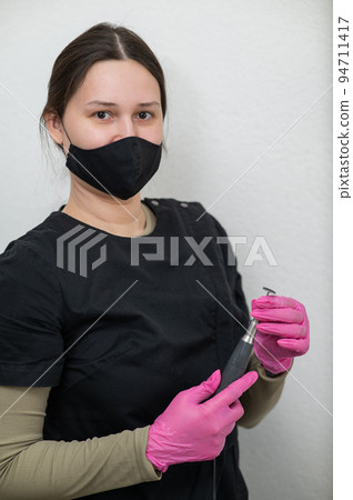 The pedicure master stands against the background of a white wall and holds an apparatus with an abrasive disc. The pedicure master stands against the background of a white wall and holds an apparatus with an abrasive disc. 94711417