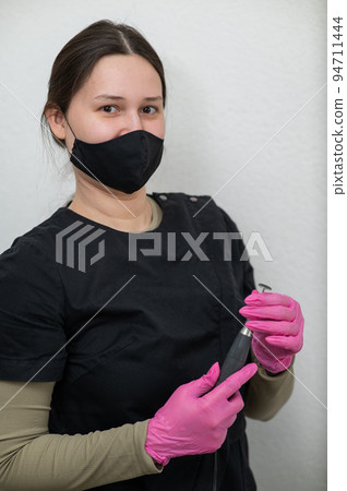 The pedicure master stands against the background of a white wall and holds an apparatus with an abrasive disc.  94711444