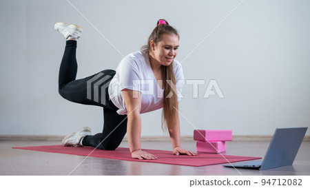 Young woman doing sports exercises at home. The girl is engaged in fitness at home on the mat and watches a training video on a laptop 94712082