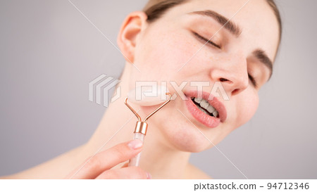 Close-up portrait of a woman using a quartz roller massager on her cheek for an alternative anti-aging. 94712346